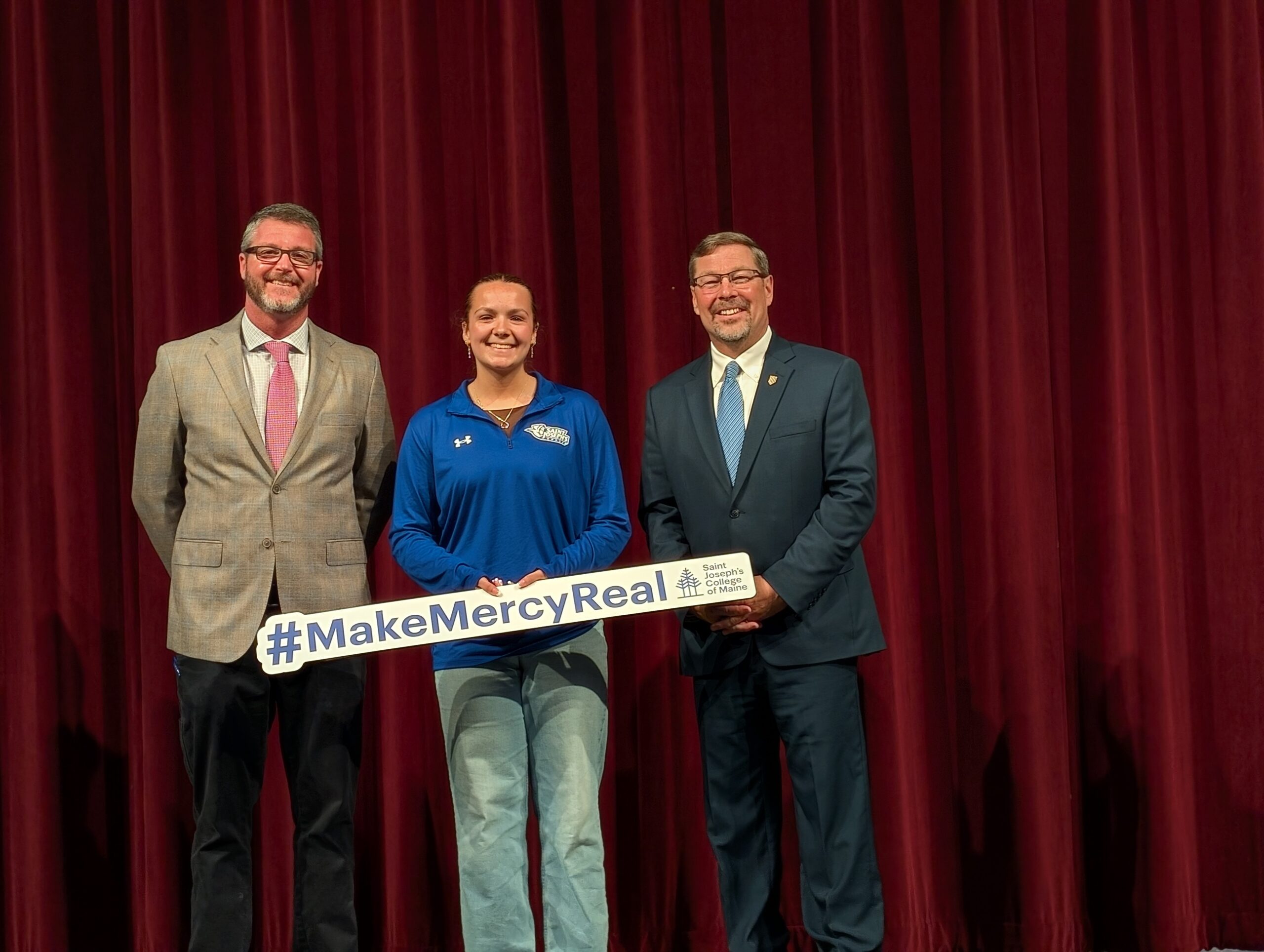 Three people stand in front of a red curtain. #MakeMercyReal Winner Adriana Pettengill, in a blue pullover, holds a #MakeMercyReal sign while the two people beside her, both in suits, celebrate her full ride achievement.