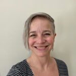 A person with short, light gray hair and a side part smiles at the camera. Wearing a black and white striped shirt, they stand in front of a plain, light-colored wall—one of SJC New Employees joining in January 2026.