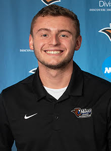 A young man with short light brown hair smiles at the camera. He is wearing a black collared shirt with an orange and blue logo on the left side, standing in front of a blue backdrop with white text and logos.