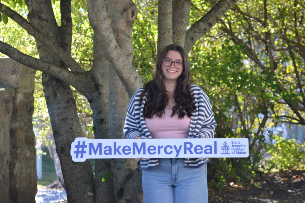 At Saint Joseph’s College of Maine, mercy is more than a founding value, it is a living legacy put into action every day. 2 A student smiling and holding a #MakeMercyReal sign at Saint Joseph’s College of Maine in front of trees, highlighting the school’s living legacy.