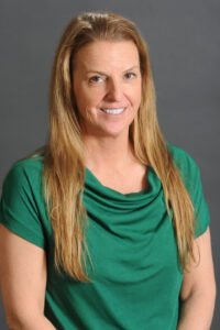 A woman with long, straight, light brown hair is smiling and wearing a green short-sleeved top, posed in front of a plain gray background, reflecting a warm and welcoming atmosphere ideal for a Counseling Center focused on mental health.