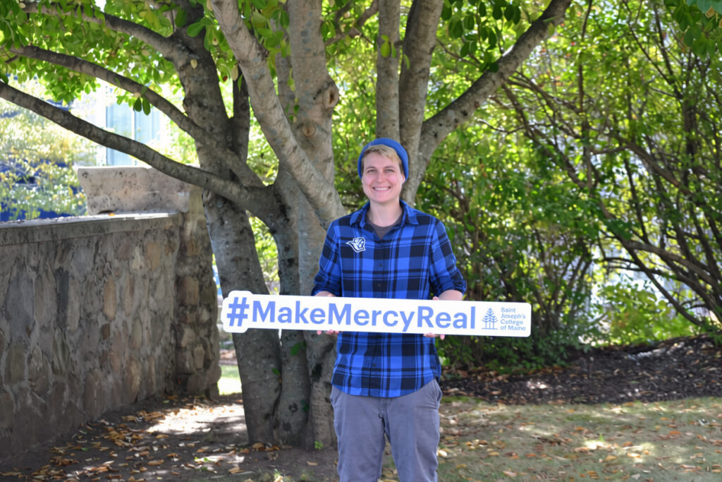 At Saint Joseph’s College of Maine, mercy is more than a founding value, it is a living legacy put into action every day. 2 Person in a blue plaid shirt and beanie stands outside under a tree, smiling and holding a sign that says #MakeMercyReal with the Sisters of Mercy of the Americas logo, embodying the founding value of mercy cherished by Saint Joseph’s College of Maine.