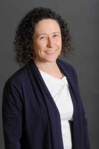 A woman with curly dark hair wearing a navy blue cardigan, white top, and brown earrings stands in front of a plain gray background, smiling at the camera, representing the Counseling Center and its mental health services.