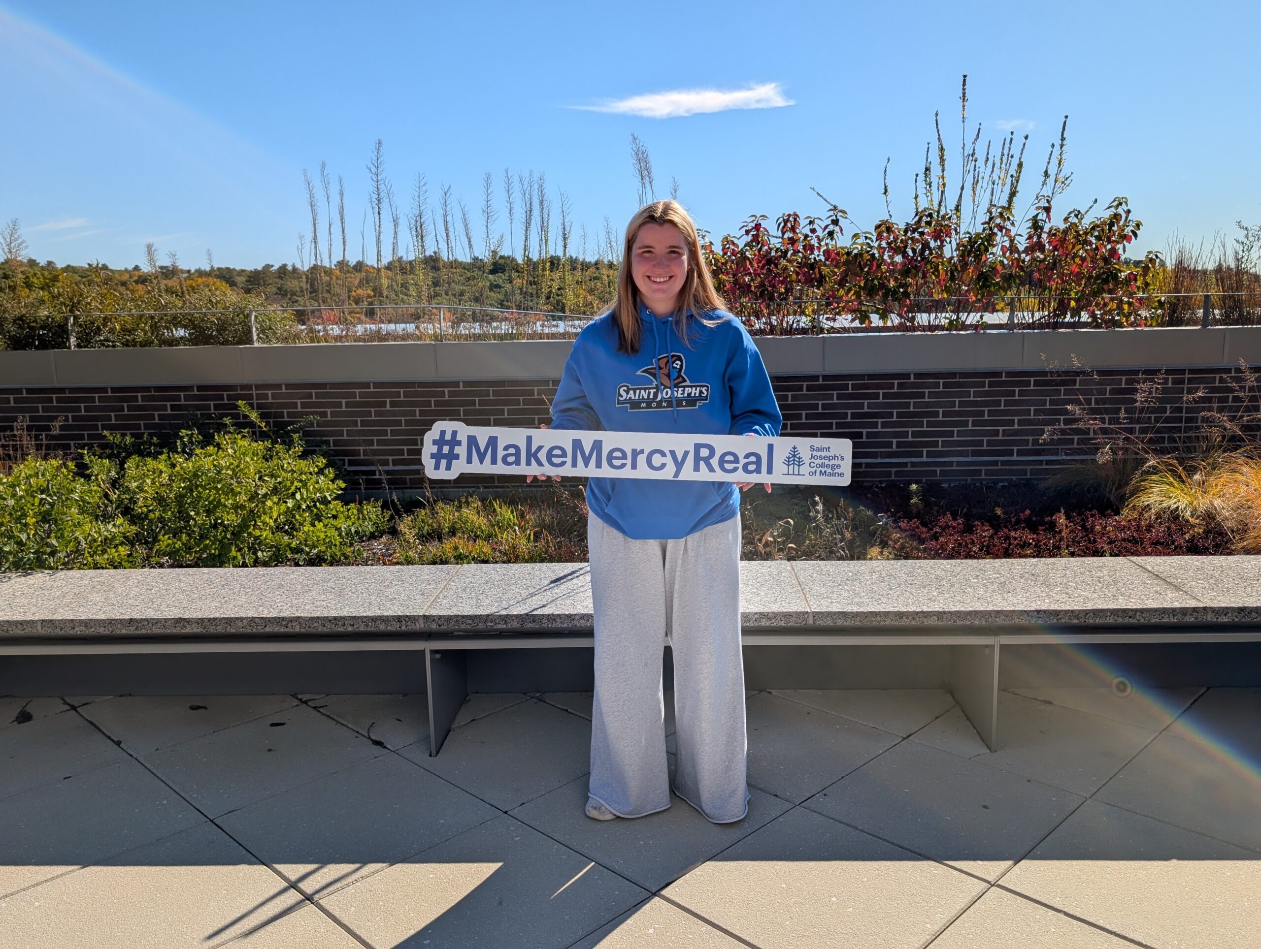 A Waltham High School student wearing a Saint Joseph’s College of Maine hoodie holds a sign that reads #MakeMercyReal along with the college’s name and logo.