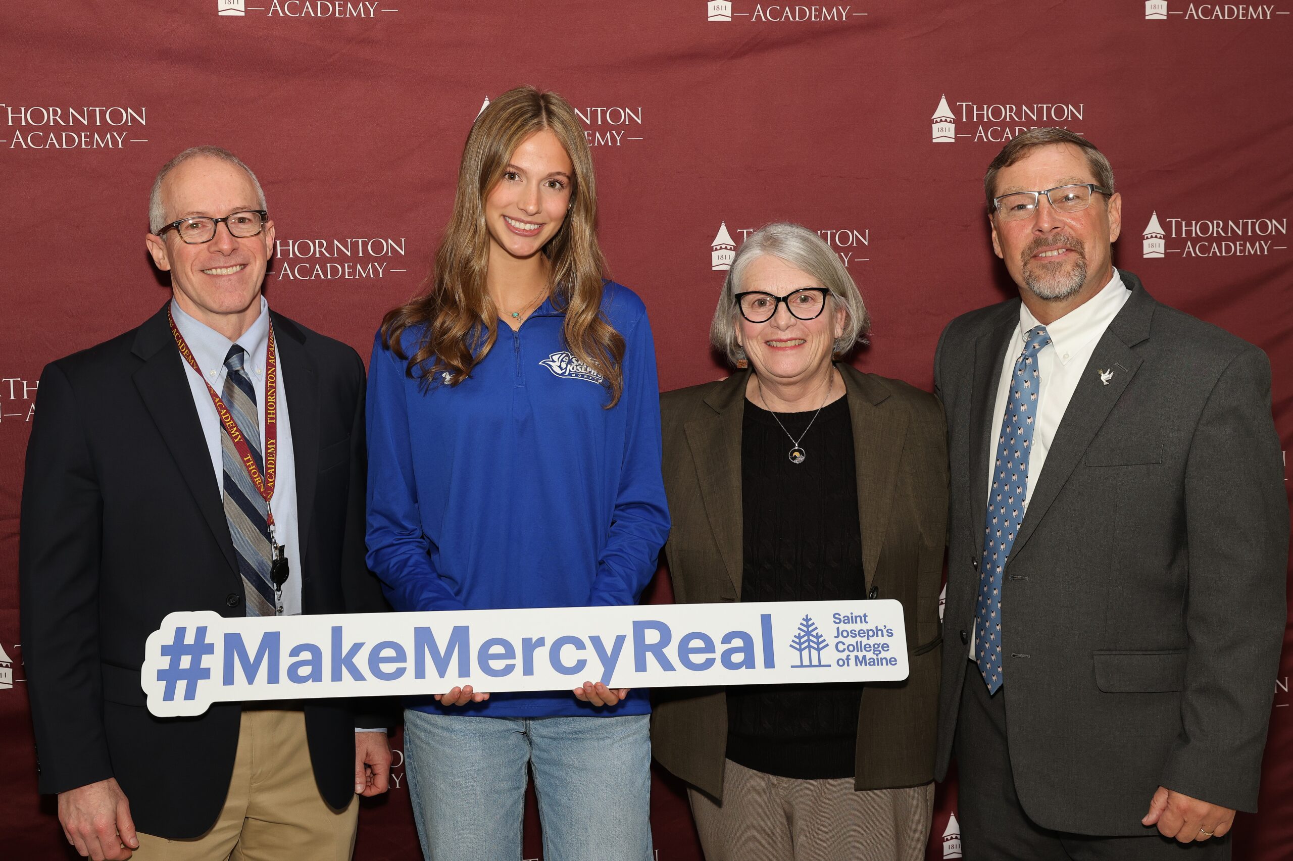 Four people stand smiling in front of a red Thornton Academy backdrop. The woman second from left, Lindsay Levesque, holds a sign reading #MakeMercyReal Saint Joseph’s College to celebrate her MakeMercyReal Scholarship.
