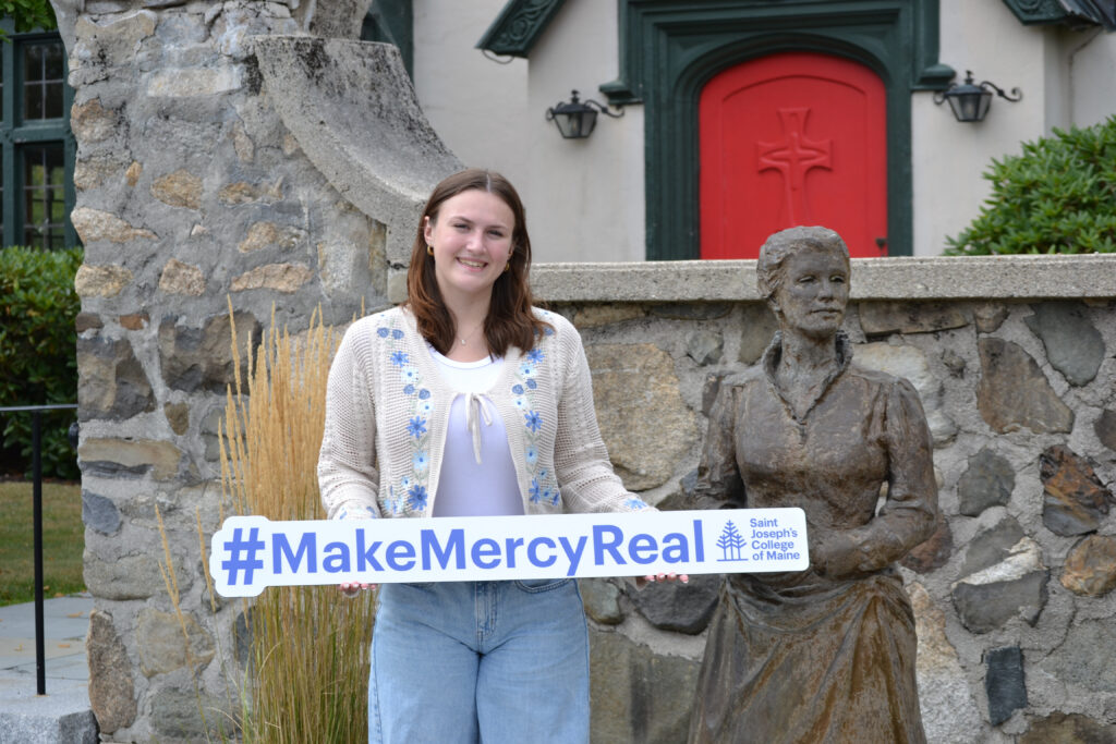 At Saint Joseph’s College of Maine, mercy is more than a founding value, it is a living legacy put into action every day. 3 A young woman stands next to a statue, holding a sign that reads #MakeMercyReal with the Saint Joseph’s College of Maine logo, embodying the founding value of mercy in front of a stone wall and building.