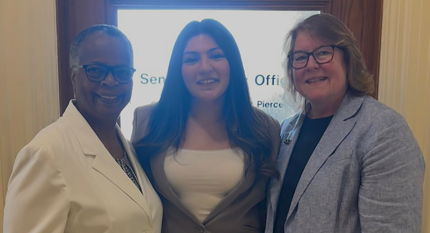 Three women stand together smiling in front of a wooden door, dressed in business attire. The Saint Josephs College of Maine logo is in the lower right corner.