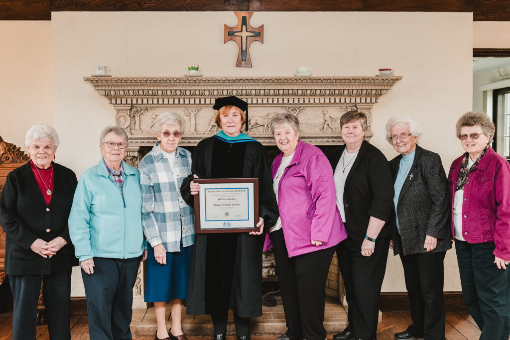 A Legacy of Mercy: Saint Joseph’s College Celebrates Commencement 2026 1 Eight women stand side by side indoors in front of a fireplace; one in the center, celebrating Commencement 2026 at Saint Joseph’s College, wears academic regalia and holds a framed certificate. A decorative cross hangs on the wall above them.