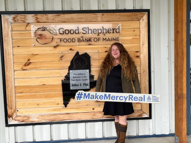 At Saint Joseph’s College of Maine, mercy is more than a founding value, it is a living legacy put into action every day. 1 A woman with long hair stands in front of the Good Shepherd Food Bank of Maine sign, holding a #MakeMercyReal sign and smiling, embodying the founding value of mercy celebrated at Saint Joseph’s College of Maine.