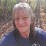 A woman with wet hair and a blue shirt smiles at the camera while standing in a sunlit forest with fallen leaves, capturing the joy of SJC New Employees in March 2026.