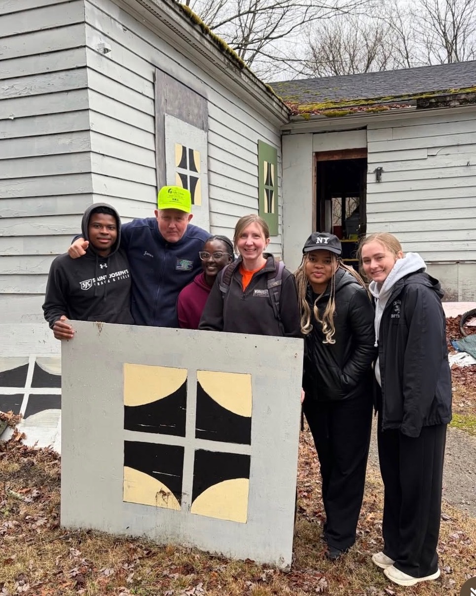 Six people stand outside a weathered, grey building, smiling at the camera. Marking a Milestone of Service, they hold a large panel painted with a black and yellow geometric pattern. Leafless trees and a door with missing glass appear in the background.