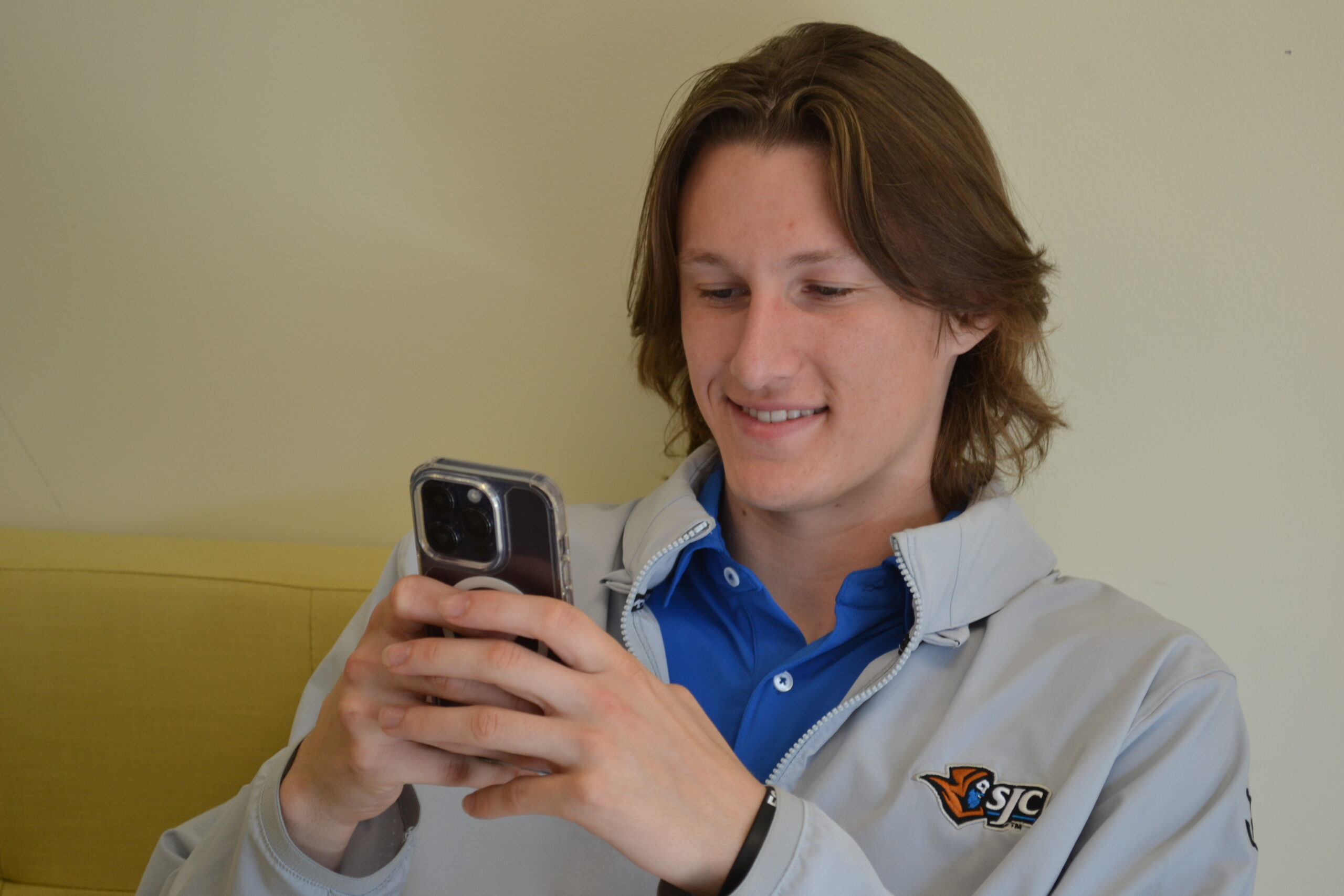 A young person with long brown hair smiles while using a smartphone, perhaps participating in the TikTok Scholarship Challenge. They wear a light gray jacket with the SJC logo and sit on a yellow couch against a plain light wall, representing Saint Joseph’s College of Maine.