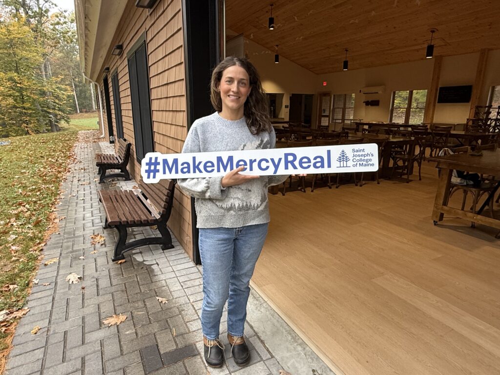 At Saint Joseph’s College of Maine, mercy is more than a founding value, it is a living legacy put into action every day. 3 A woman stands on a patio holding a sign that reads #MakeMercyReal with the Saint Joseph’s College of Maine logo, honoring the school’s commitment to mercy.