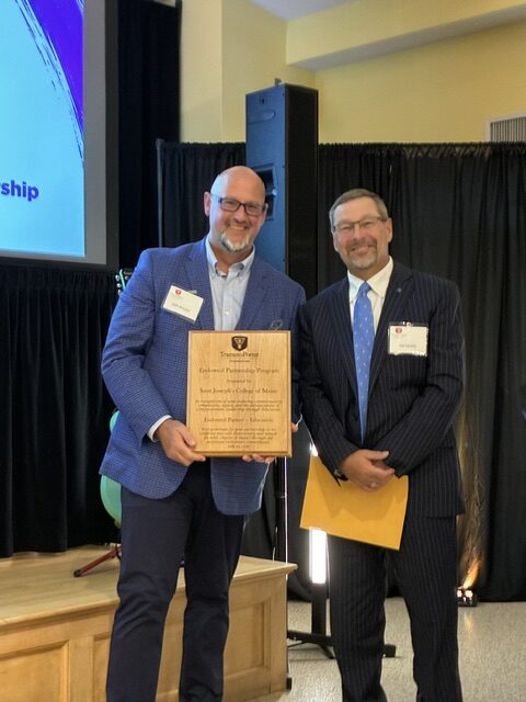 Saint Joseph’s College Leaders Celebrate TramutoPorter Foundation’s 25th Anniversary Gala 1 Two men in suits smiling, one holding a wooden award plaque. They stand on a stage with a black curtain behind them and a presentation screen partially visible to the left—capturing a memorable moment at the Saint Joseph’s College Anniversary Gala, presented by the TramutoPorter Foundation.