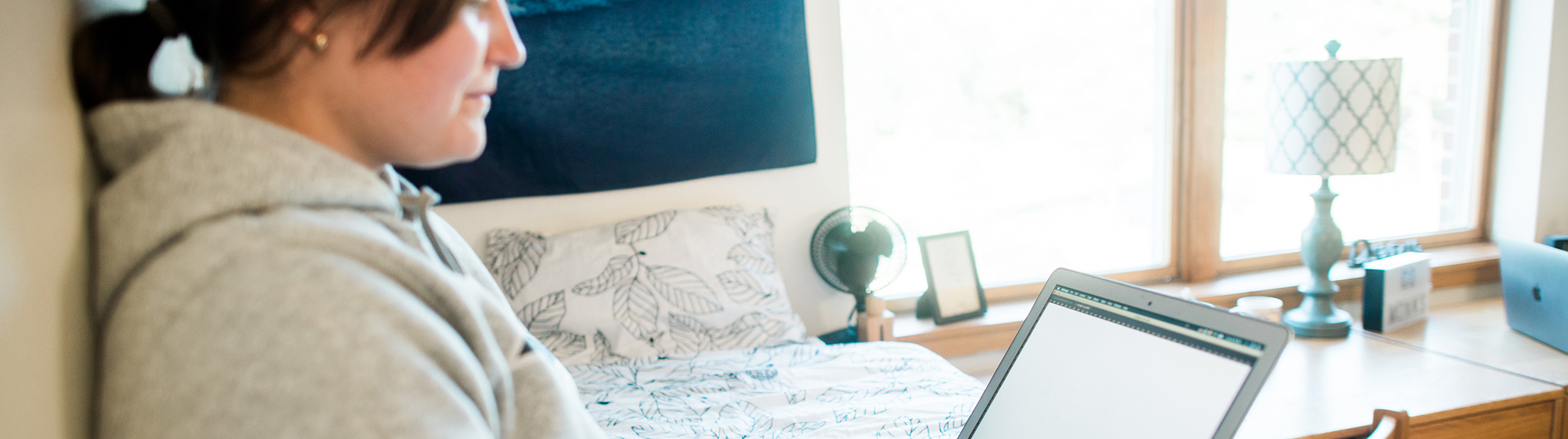A person in a gray hoodie sits in a bright bedroom holding an open laptop. The bed features patterned bedding, and natural light pours in. On the wooden windowsill are gently used home goods like a lamp, fan, and framed photo—perfect for Rest Pods.
