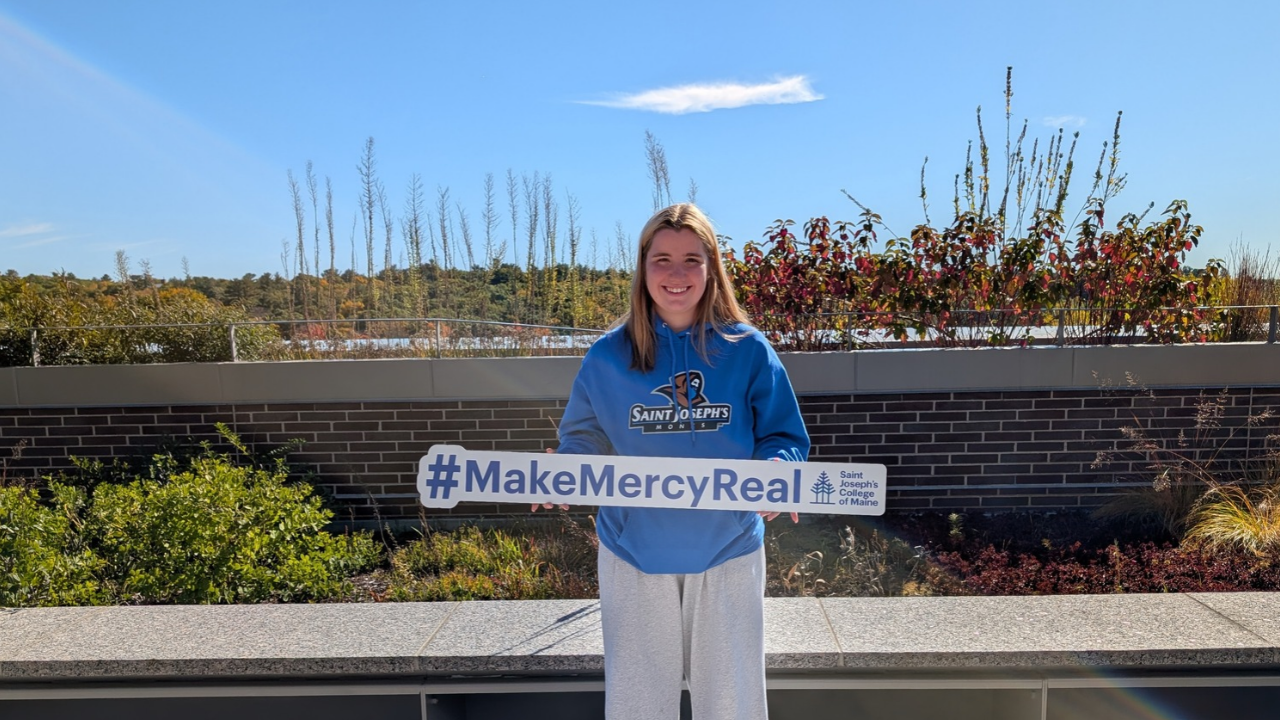 A Waltham High School student wearing a Saint Joseph’s College of Maine hoodie holds a sign that reads #MakeMercyReal.